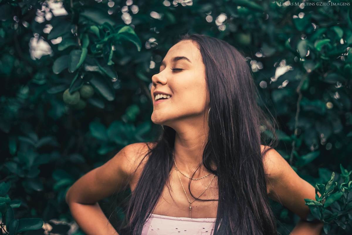 fotografia da debutante sorrindo de olhos fechados em  meio a vegetação durante ensaio fotográfico de 15 anos na cidade de Virgem da Lapa - MG