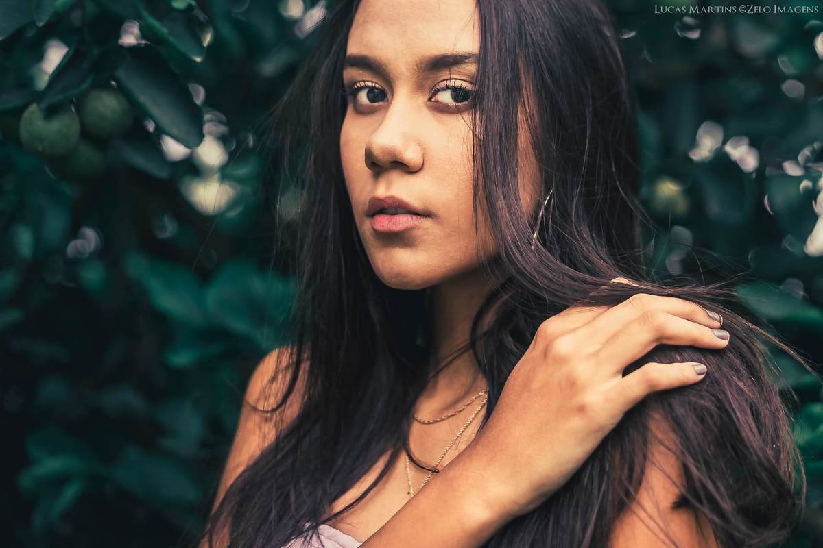 fotografia da debutante olhando para a foto, com pose com a mão no ombro, em meio a vegetação durante ensaio fotográfico 15 anos, na cidade de Virgem da Lapa, Minas Gerais