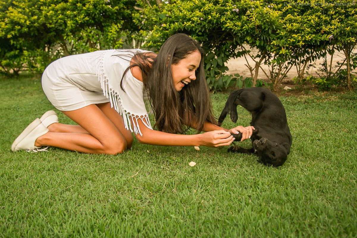 fotografia da debutante brincando com um cachorro preto em  um gramado, debutante sorrindo. Durante ensaio fotográfico de 15 anos em Virgem da Lapa, MG