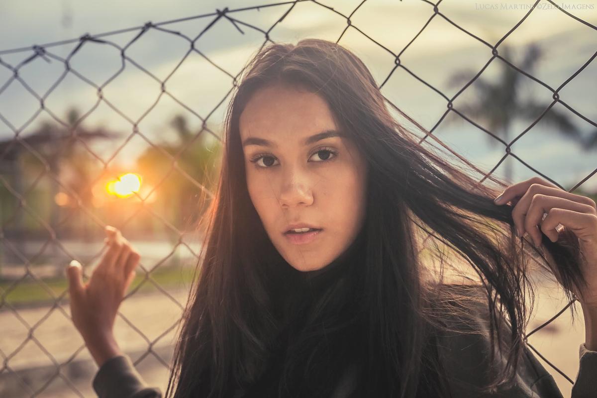 fotografia de debutante encostada na em cerca de grades mexendo no cabelo com por do sol ao fundo em ensaio fotográfico 15 anos realizado na cidade de Virgem da Lapa, Minas Gerais