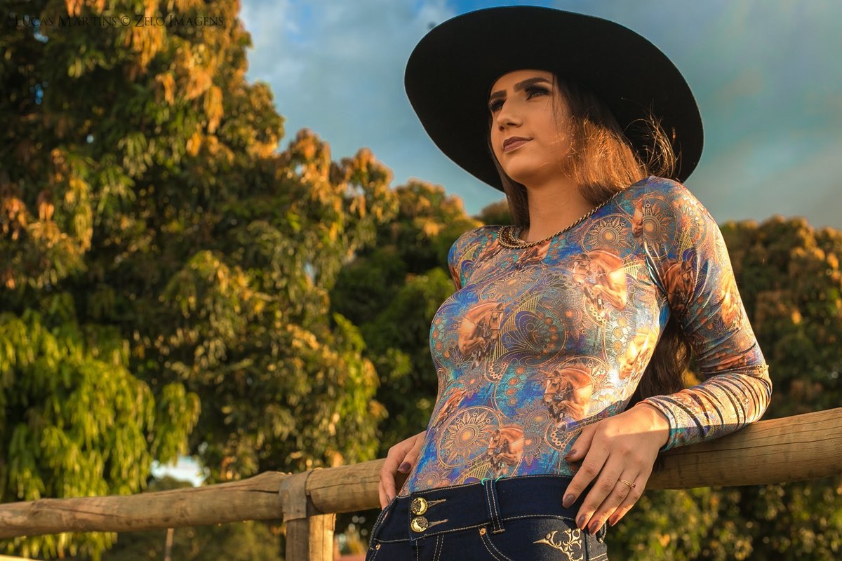 fotografia da debutante de chapéu, apoiada em cerca de madeira e no fundo as copas de mangueiras, durante ensaio fotográfico 15 anos em Araçuaí, Minas Gerais 