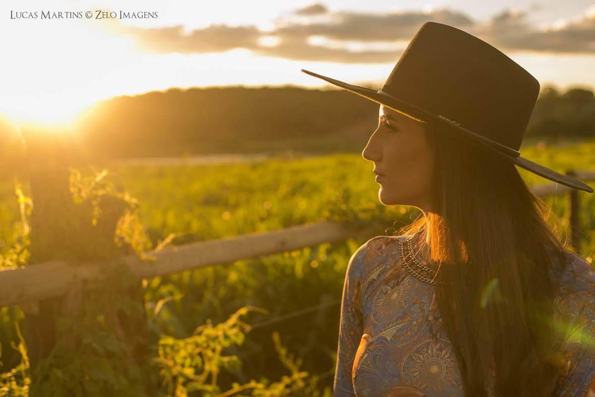 Fotografia da debutante de chapéu preto, observando a paisagem durante o por do sol em haras, em ensaio fotográfico de 15 anos feito na cidade de Araçuaí, Minas Gerais