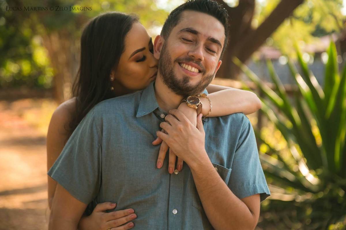 Fotografia da noiva dando um beijo no pescoço do noivo enquanto ele sorri. Em ensaio fotográfico pré casamento realizado na cidade de Berilo, Minas Gerais