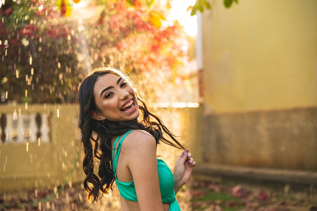 fotografia criativa - debutante sorrindo posando em frente o por do sol refletindo agua jogada pela madrinha com magueira