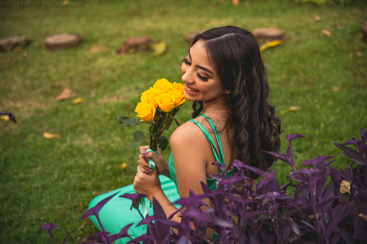 debutante posa perto da grama e de flores roxas segurando um boquet amarelo