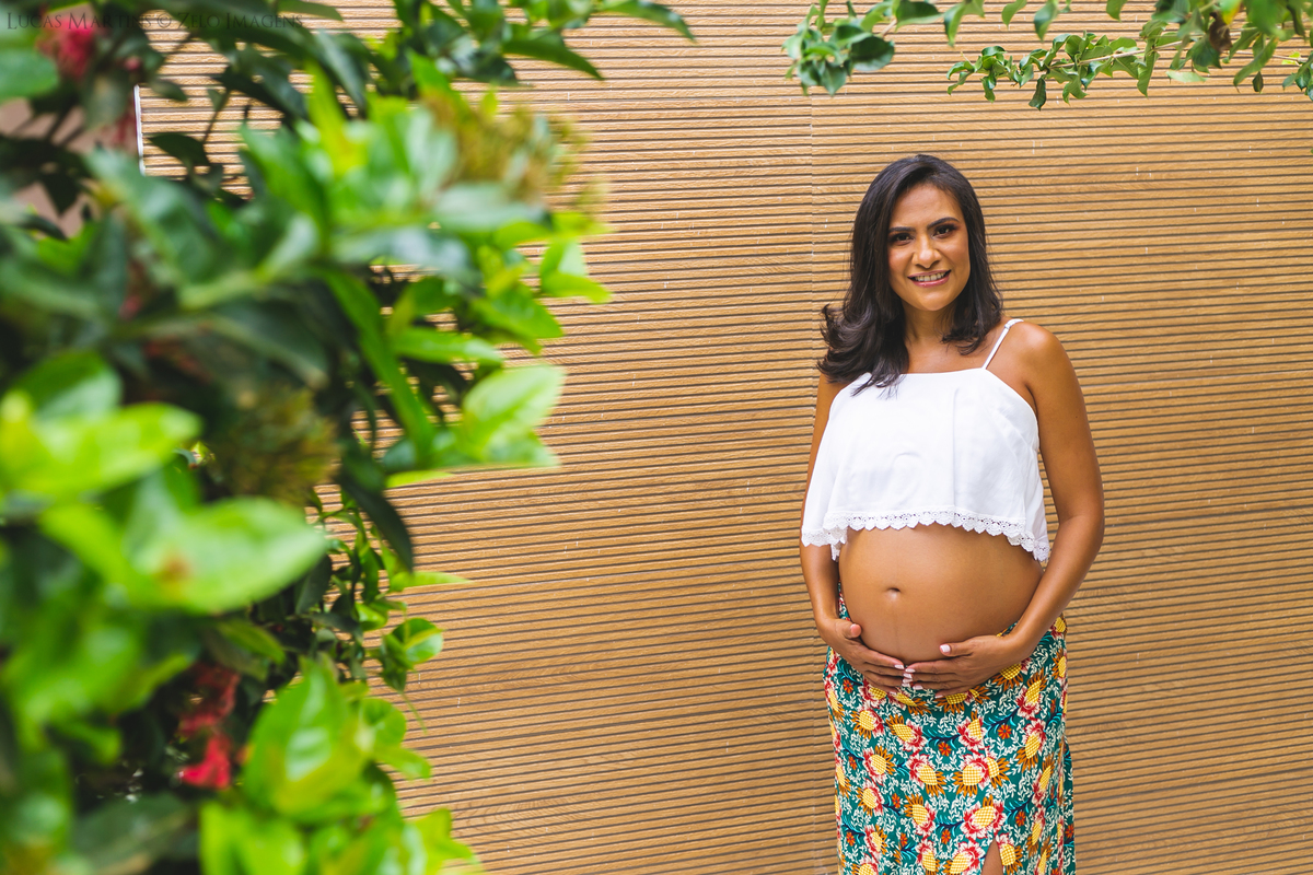 Gestante posa sorrindo ao fundo de um painel de madeira somada a moldura natural de uma planta
