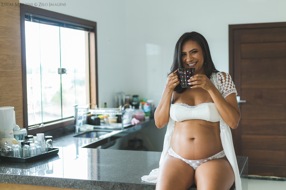 Gestante posa na bancada preta da cozinha enquanto delicia um café