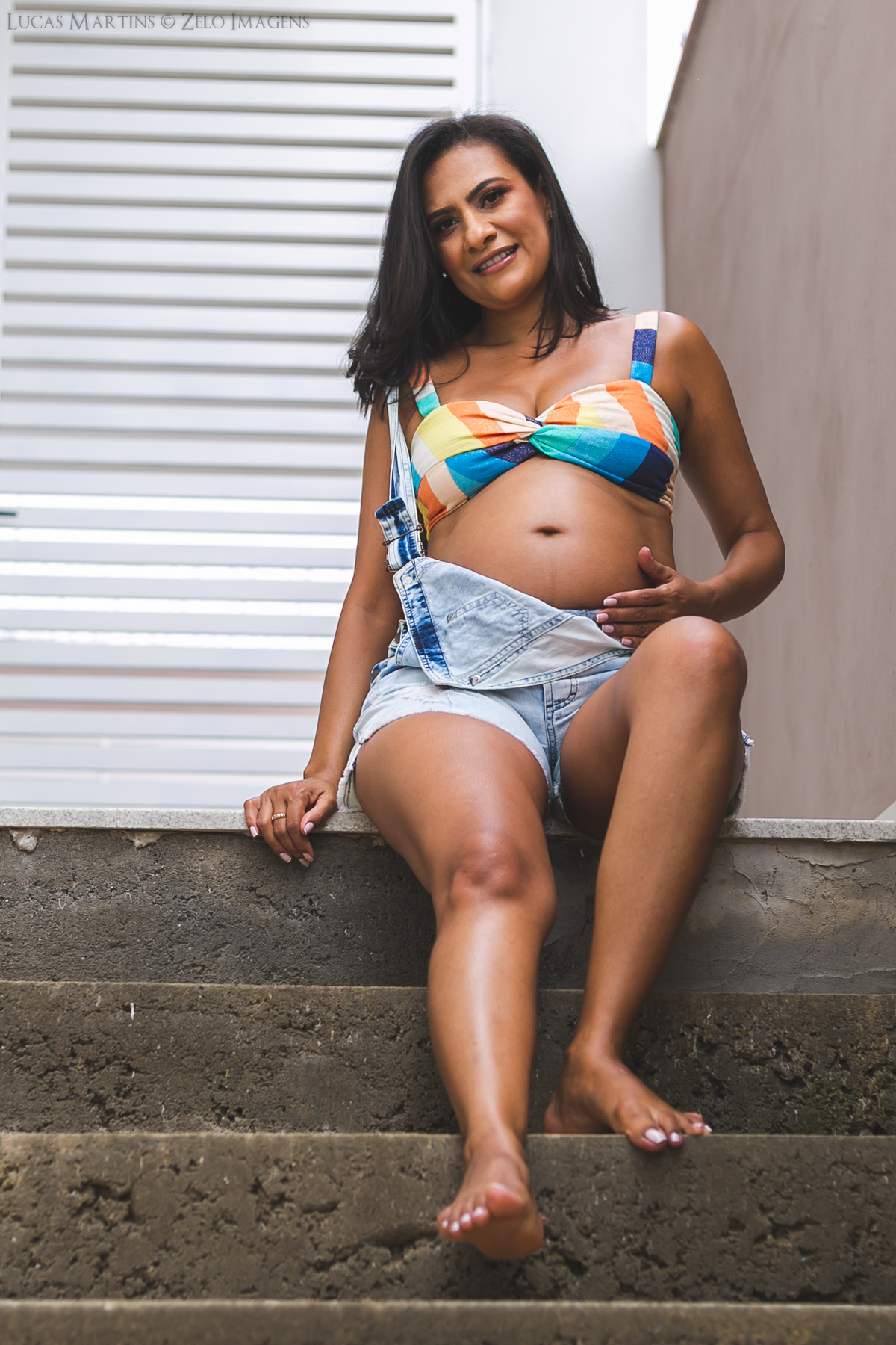Gestante posa com um look mais casual, jardineira jeans e top colorido sentada na escada de cimento