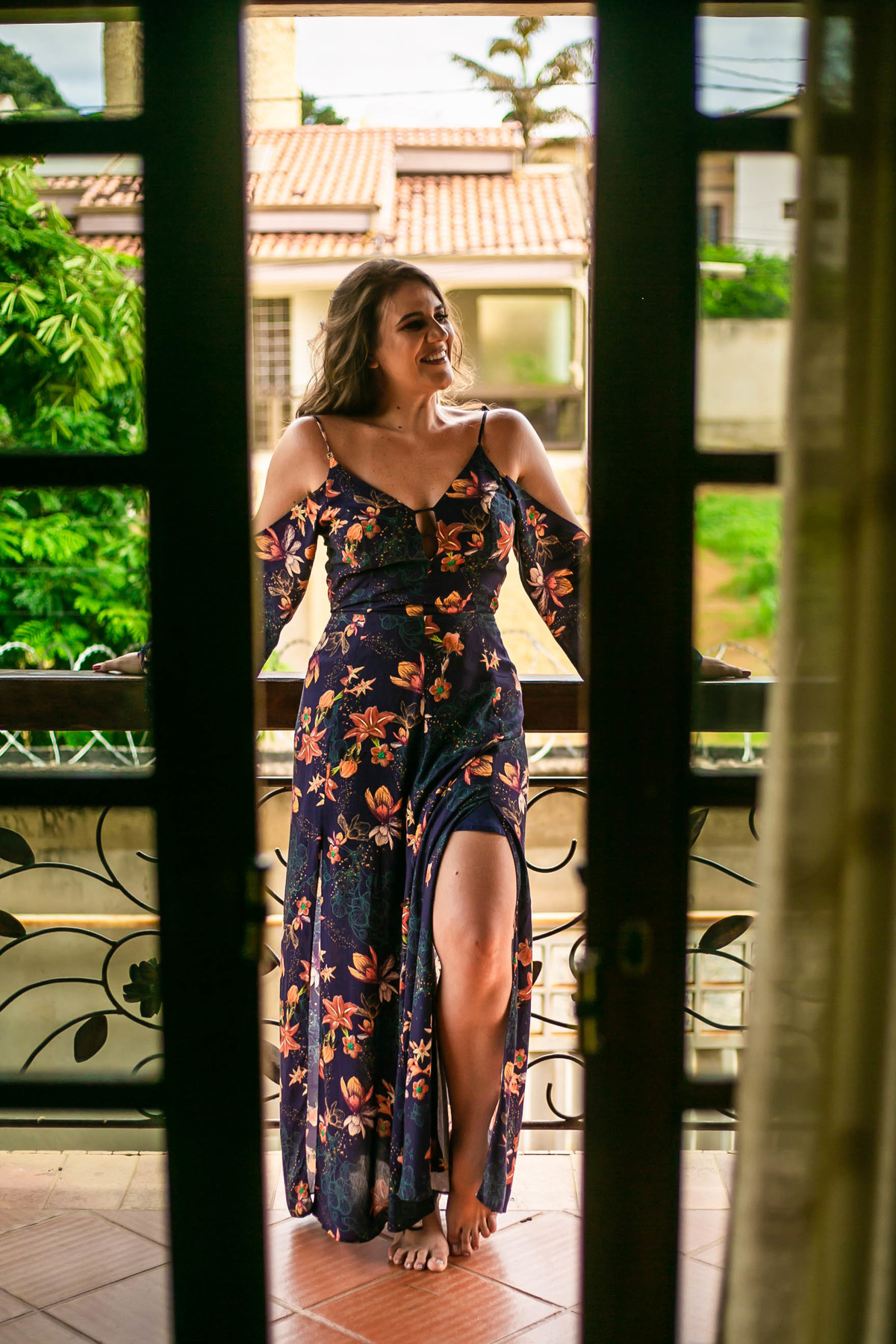 Modelo usando vestido longo azul florido, encostada numa sacada de madeira.