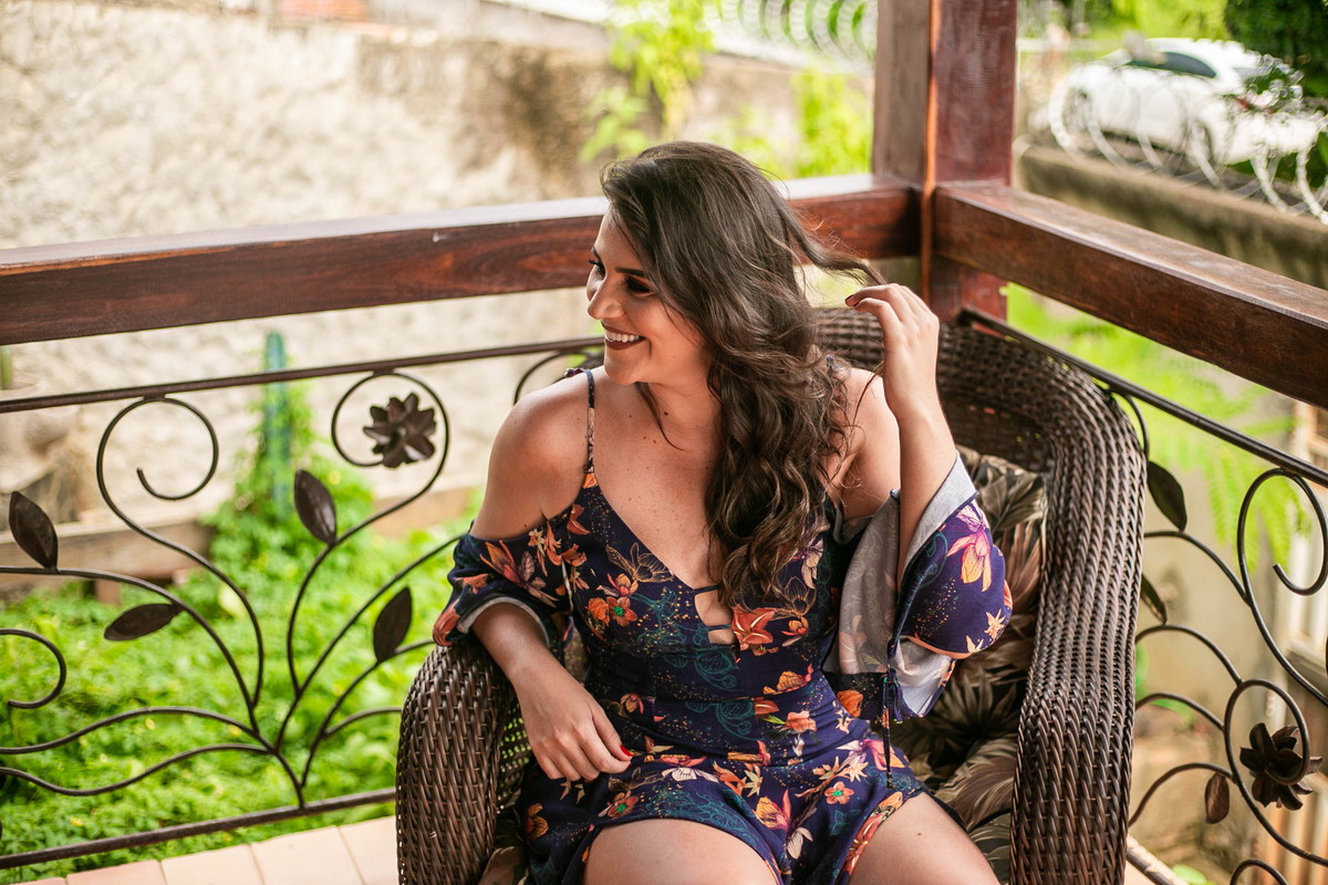 Modelo usando vestido longo azul florido, sentada numa poltrona de vime trançado.