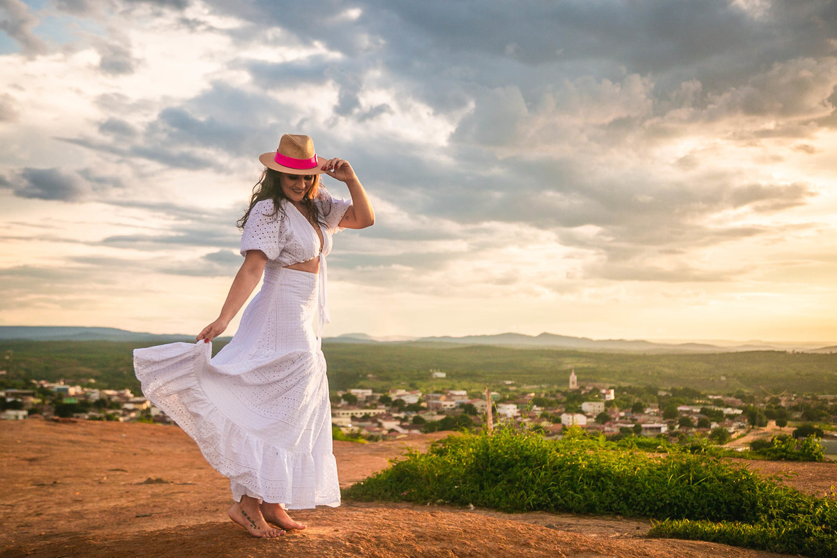 Modelo usando blusa e saia brancas e chapéu de palha ao pôr do sol.