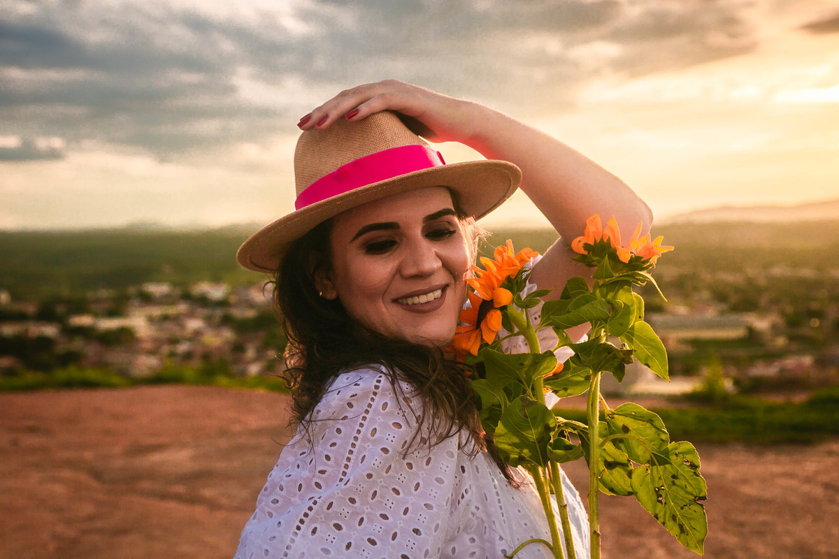 Modelo usando blusa e saia brancas e chapéu de palha, segurando um buquê de girassóis ao pôr do sol.