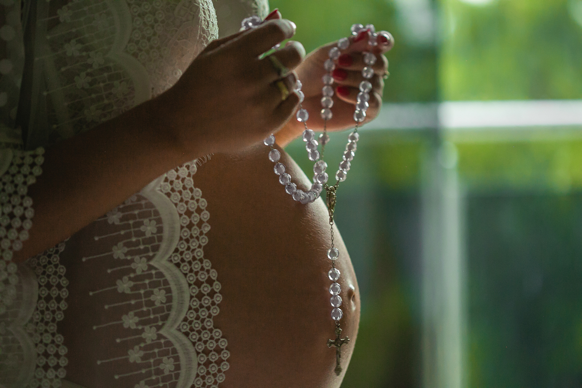 Foto em close na barriga da gestante, que tem em suas mãos um terço de contas transparentes. 