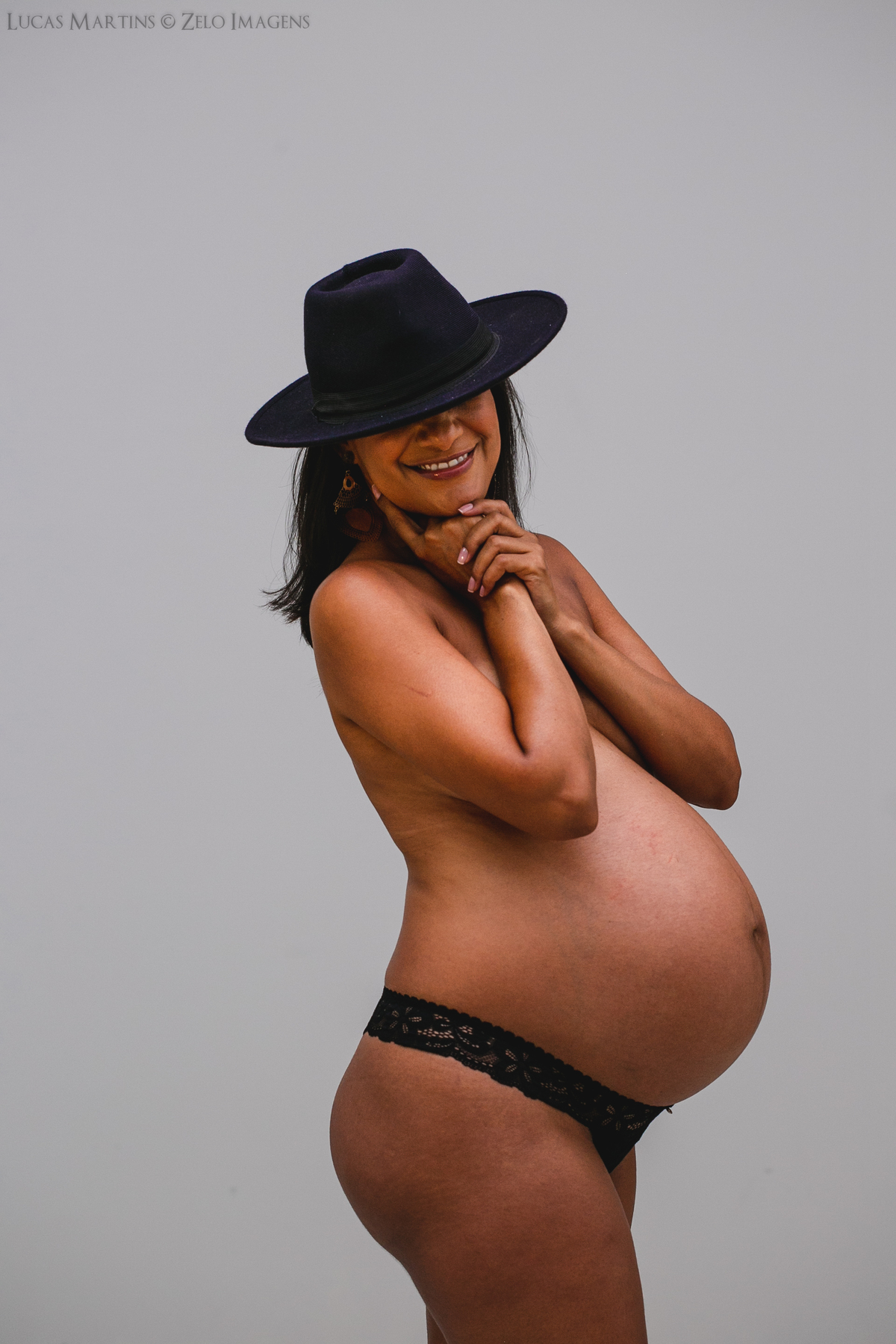 Gestante em estúdio, usando apenas chapéu e calcinha pretos. Ela posa de perfil coma as mãos proximas ao rosto e o chapeu cobrindo os olhos.