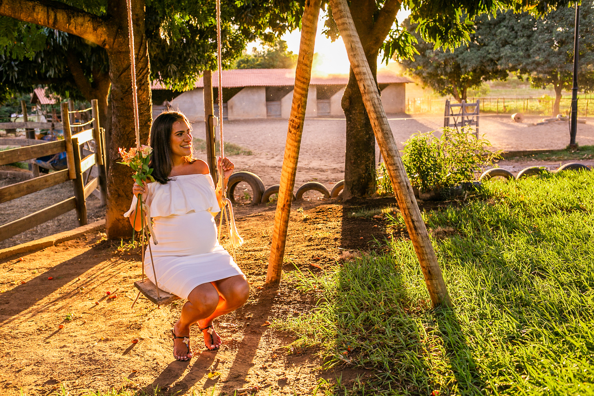 Gestando sentada em gangorra com bases de eucalipto. Ela usa vestido branco, ombro a ombro e segura um pequeno ramo de flores amarelas em sua mão.