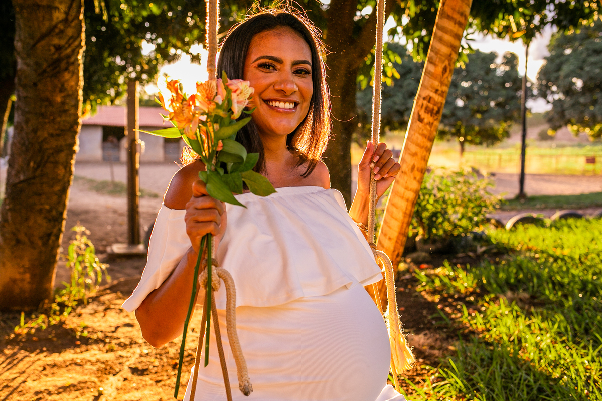 Gestando sentada em gangorra com. Ela usa vestido branco ombro a ombro e segura um pequeno ramo de flores amarelas em sua mão.
