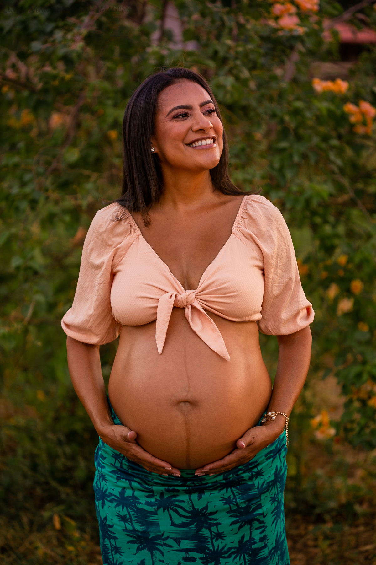 Gestante posa com cropped salmão e saia verde, apoiando levemente suas mãos abaixo da barriga.