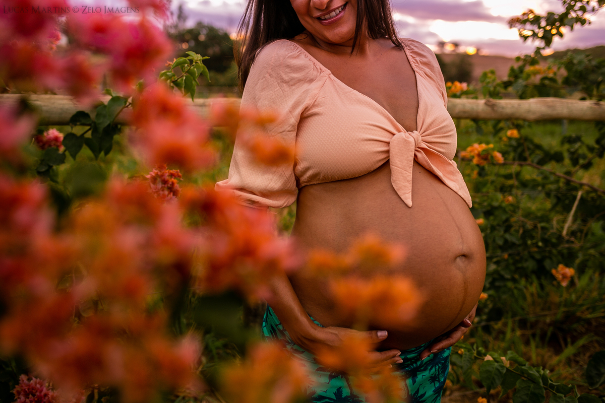 Gestante usando cropped salmão e saia longa verde, posa ligeiramente de lado apoiando suas mãos abaixo da barriga.
