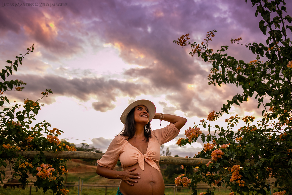 Gestante usa chapéu claro e cropped salmão. 
Posa com uma das mãos na cabeça, a outra apoiada na barriga, em frente a uma cerca de madeira e em meio a galhos floridos.