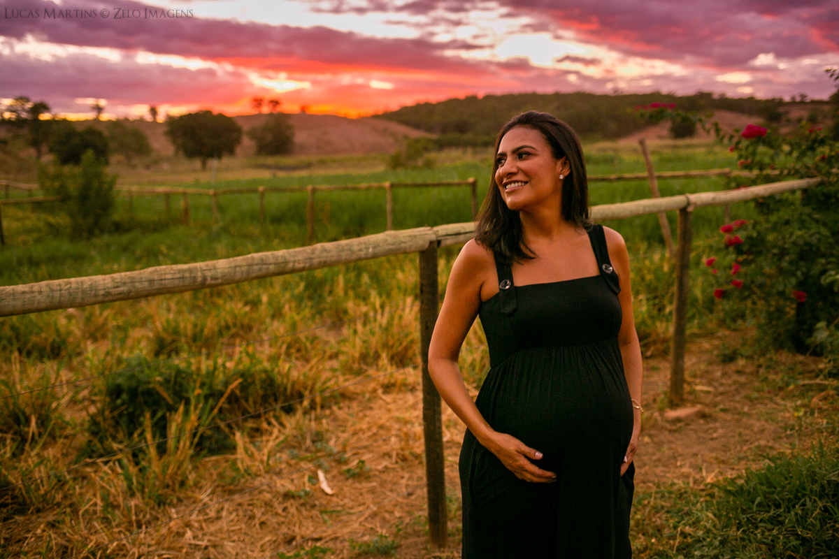 Gestante usando vestido preto posa em frente a uma cerca de madeira. Ao fundo vê-se o por do sol.