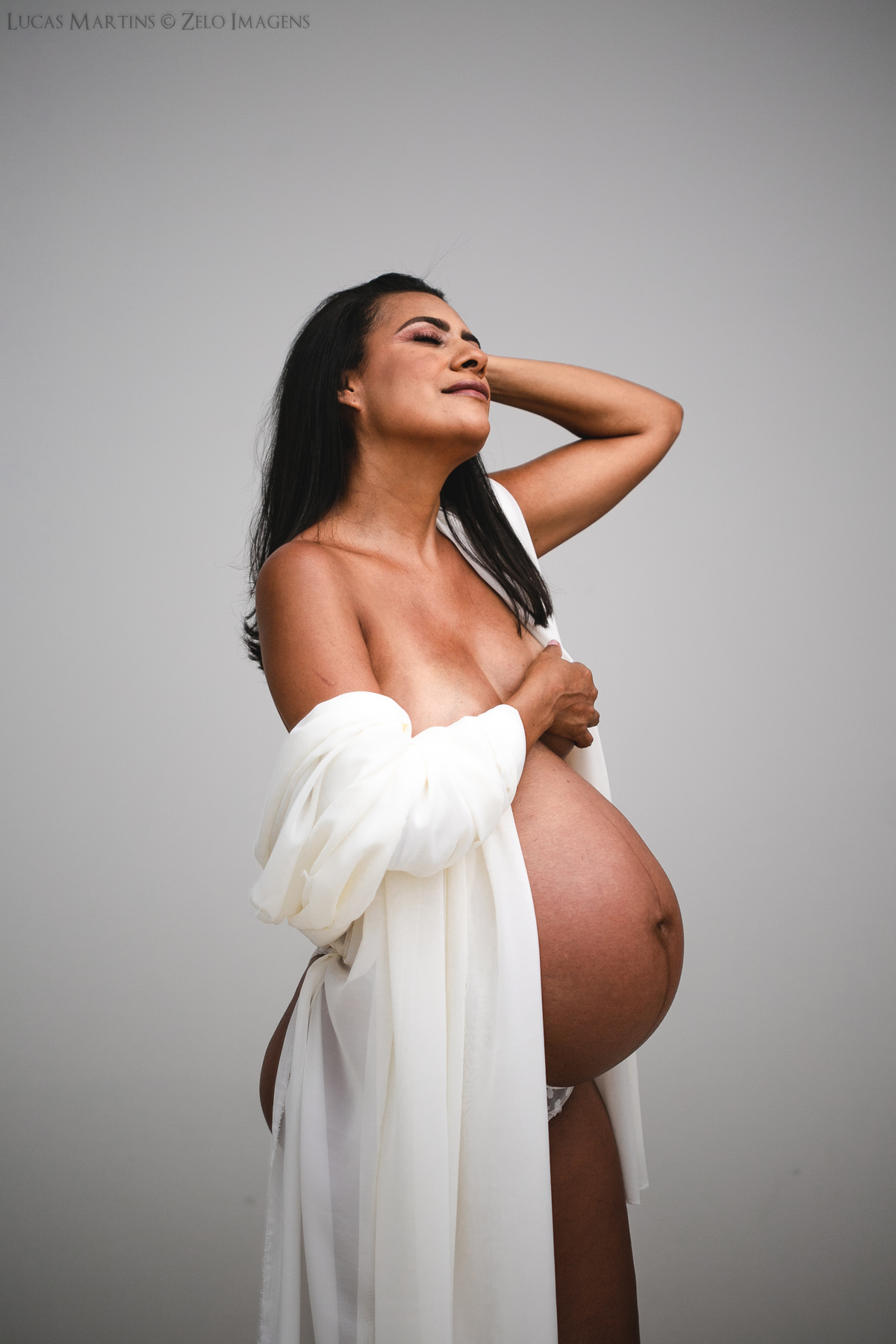 Foto em estúdio de gestante de perfil usando um tecido jogado sobre os braços.