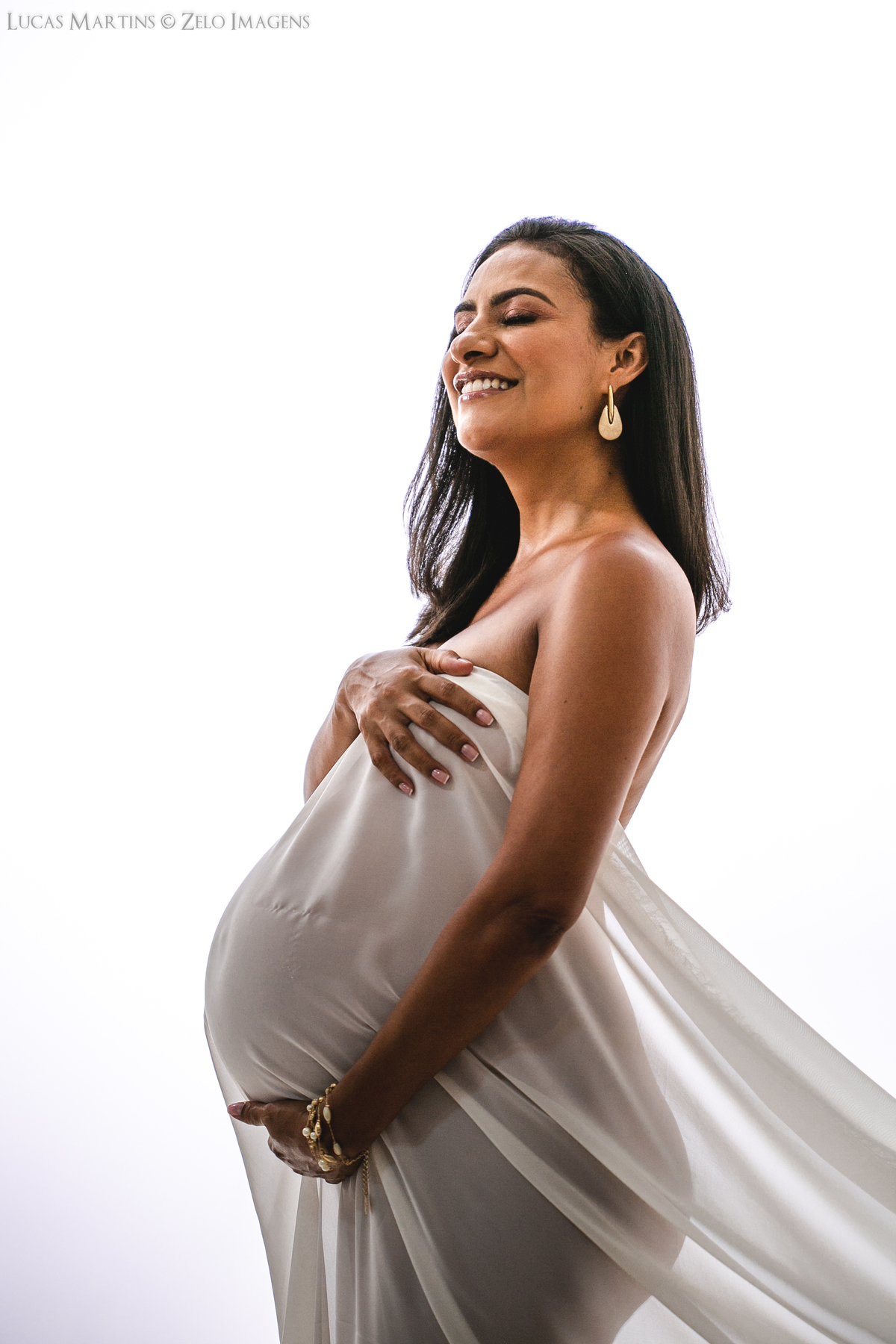 Foto em estúdio de gestante envolta em tecido branco, de perfil, evidenciando sua barriga.