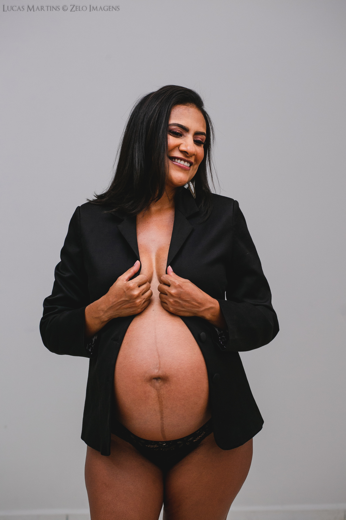 Foto em estudio de gestante trajando blazer preto aberto, cobrindo os seios, mas deixando à mostra sua barriga.