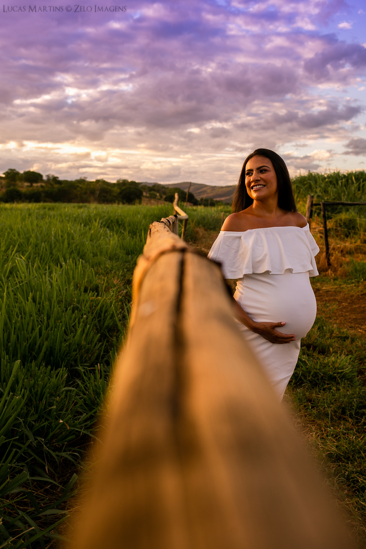 Gestante usando vestido ombro a ombro branco, próxima à cerca em campo esverdeado.