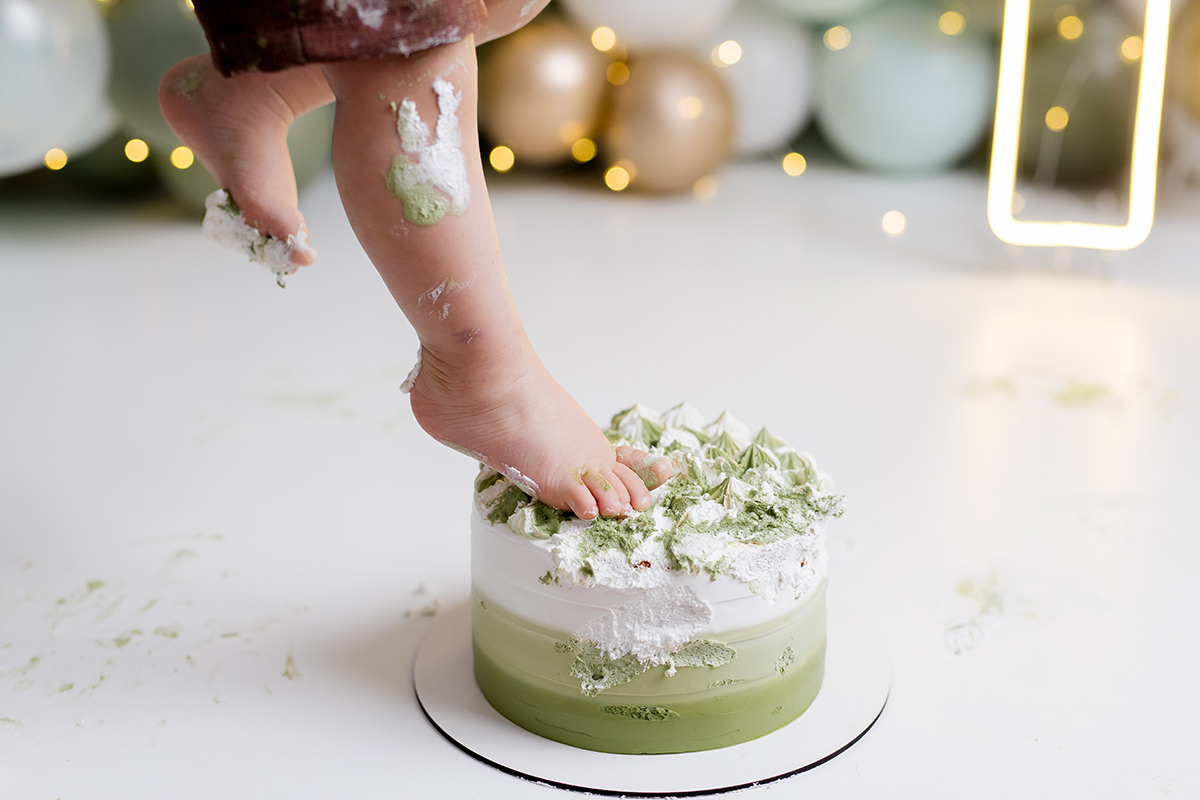 bebê encostando o pé para o bolo   com as mãos sujas no ensaio smash the cake com balõess verde, branco e dourado