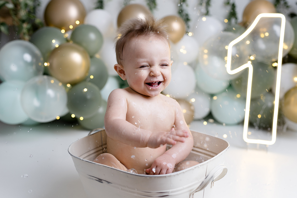 bebê sentado dentro da tina com agua ensaio smash the cake com balõess verde, branco e dourado