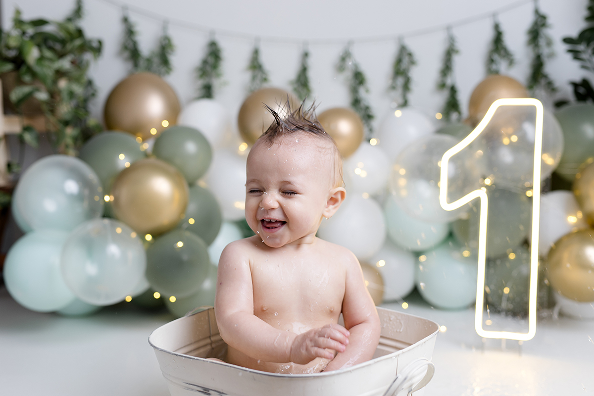 bebê sentado dentro da tina com agua ensaio smash the cake com balõess verde, branco e dourado