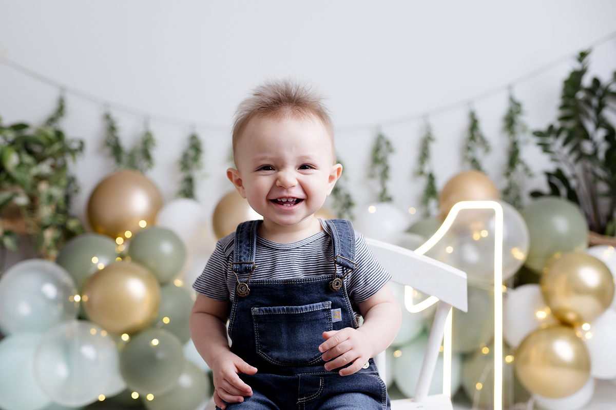 Bebê sentado na cadeira no ensaio  smash the cake com balõess verde, branco e dourado