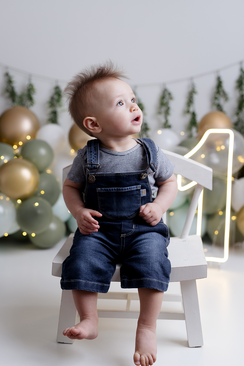 Bebê sentado na cadeira no ensaio  smash the cake com balõess verde, branco e dourado