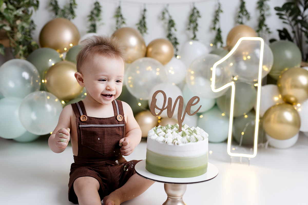 Bebê sentado olhando para o bolo no ensaio  smash the cake com balõess verde, branco e dourado