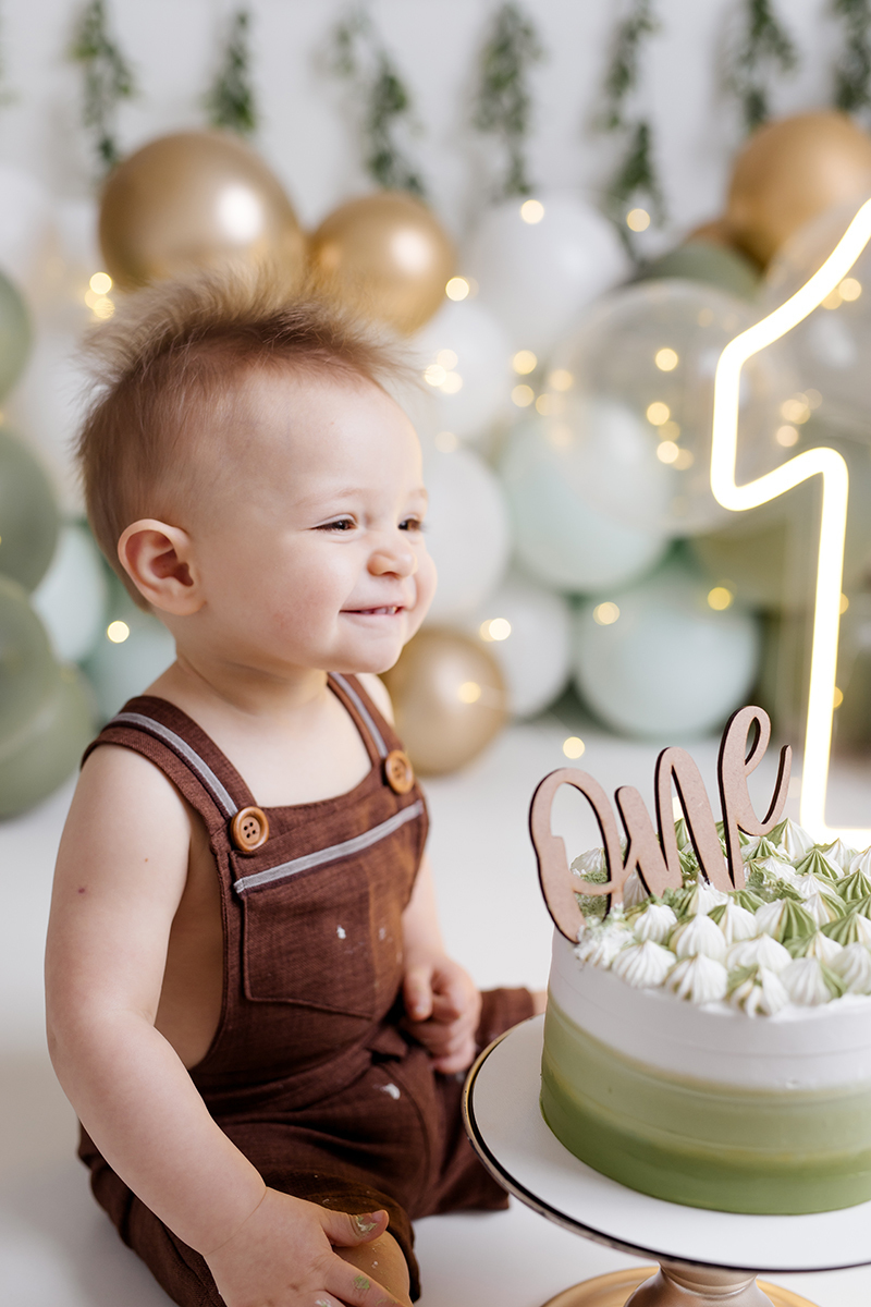 bebê sentado olhando para o bolo no ensaio  smash the cake com balõess verde, branco e dourado
