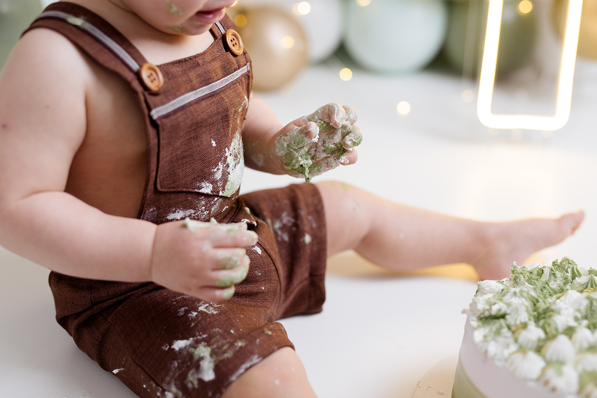bebê sentado olhando para o bolo no ensaio   com as mãos sujas no ensaio smash the cake com balõess verde, branco e dourado