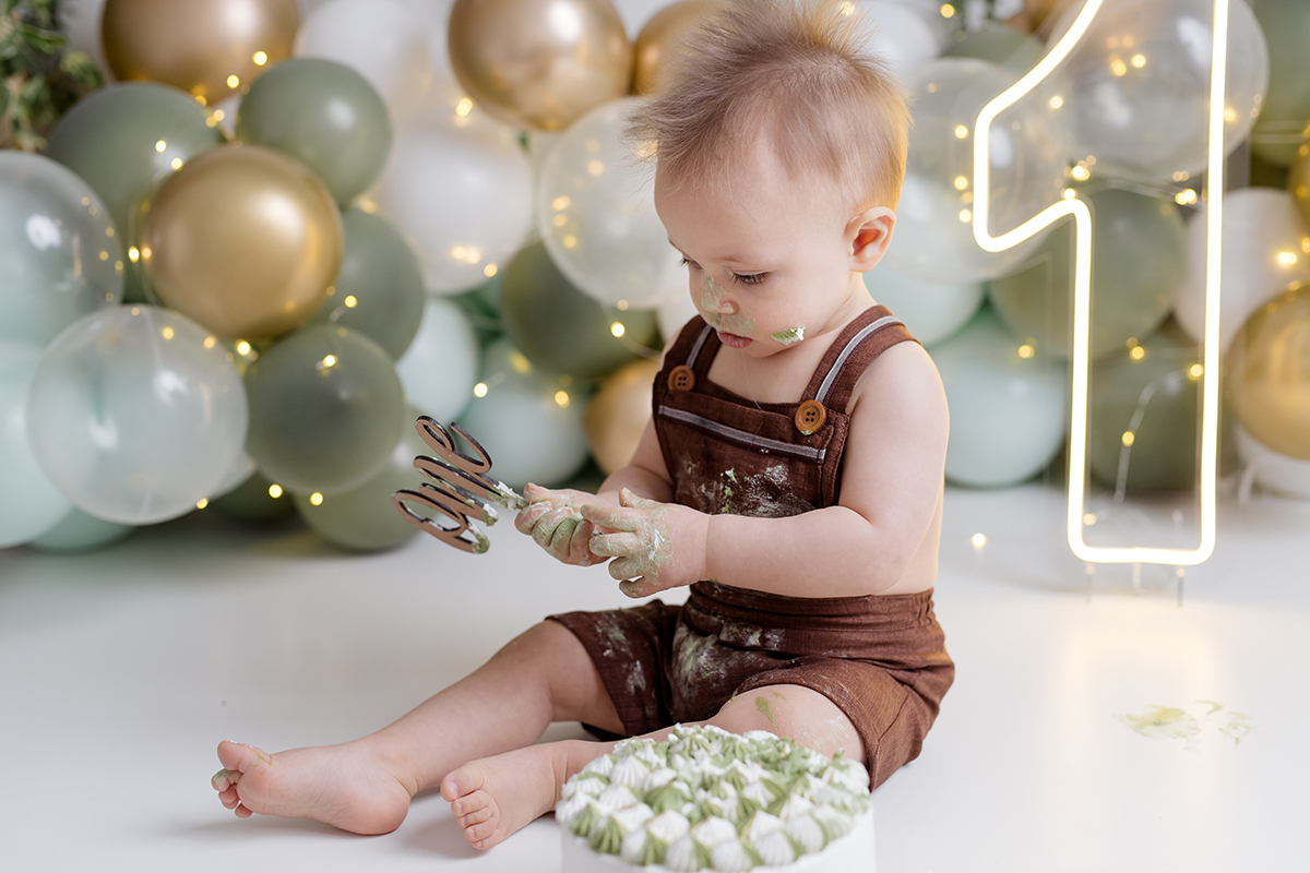 bebê sentado olhando para o bolo no ensaio   com as mãos sujas no ensaio smash the cake com balõess verde, branco e dourado