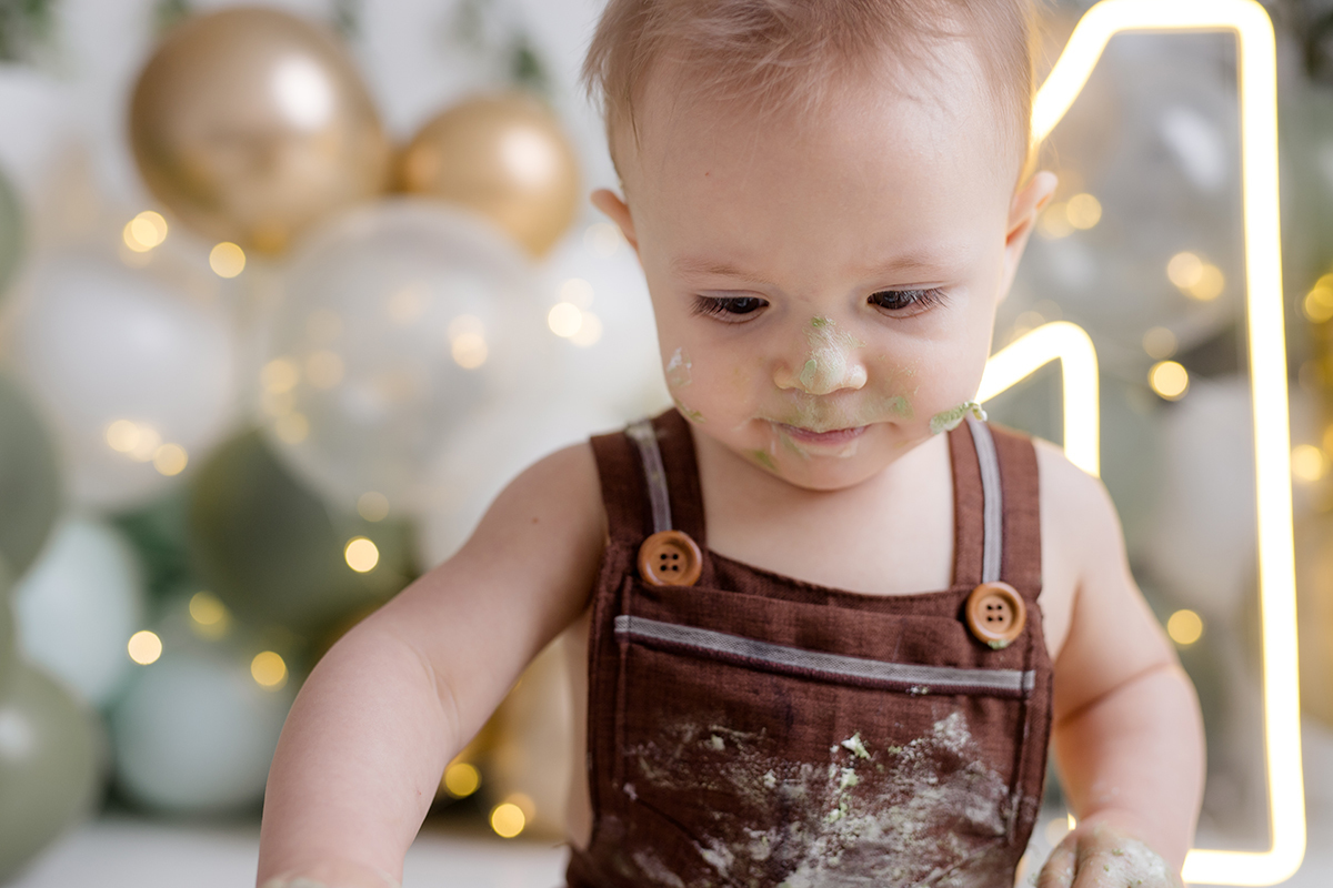 bebê sentado olhando para o bolo   com as mãos sujas no ensaio smash the cake com balõess verde, branco e dourado