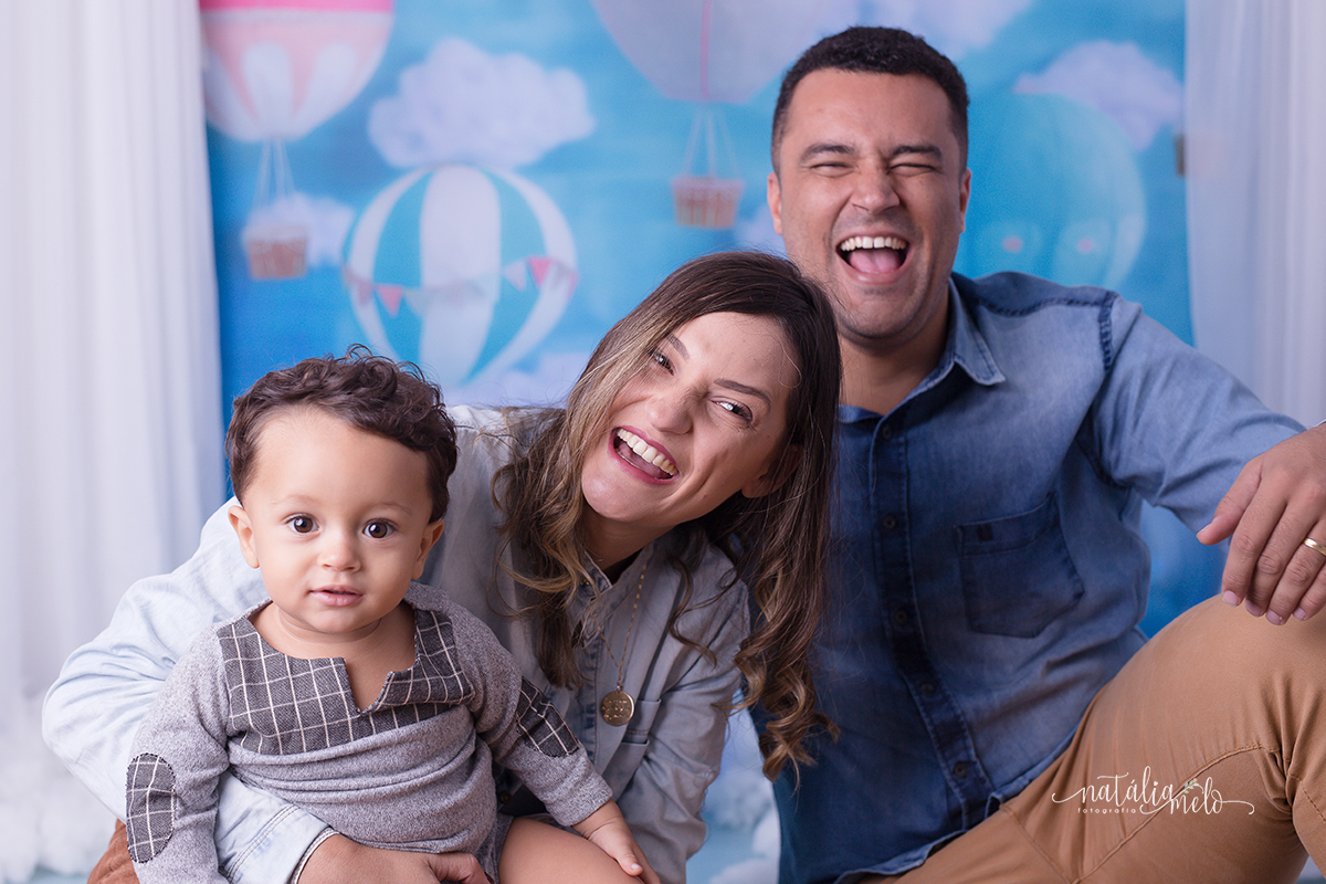 Pai e mãe abraçados com filho, rindo muito, num cenário de balões no Estúdio Natália Melo Fotografia em Belo Horizonte -