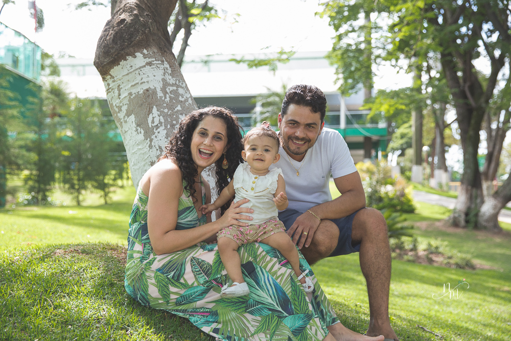 Ensaio de Família feito por Natália Melo Fotografia no Clube Jaraguá em Belo Horizonte - MG, onde pai e mãe carregam Lara e brincam com ela no colo sentados no gramad