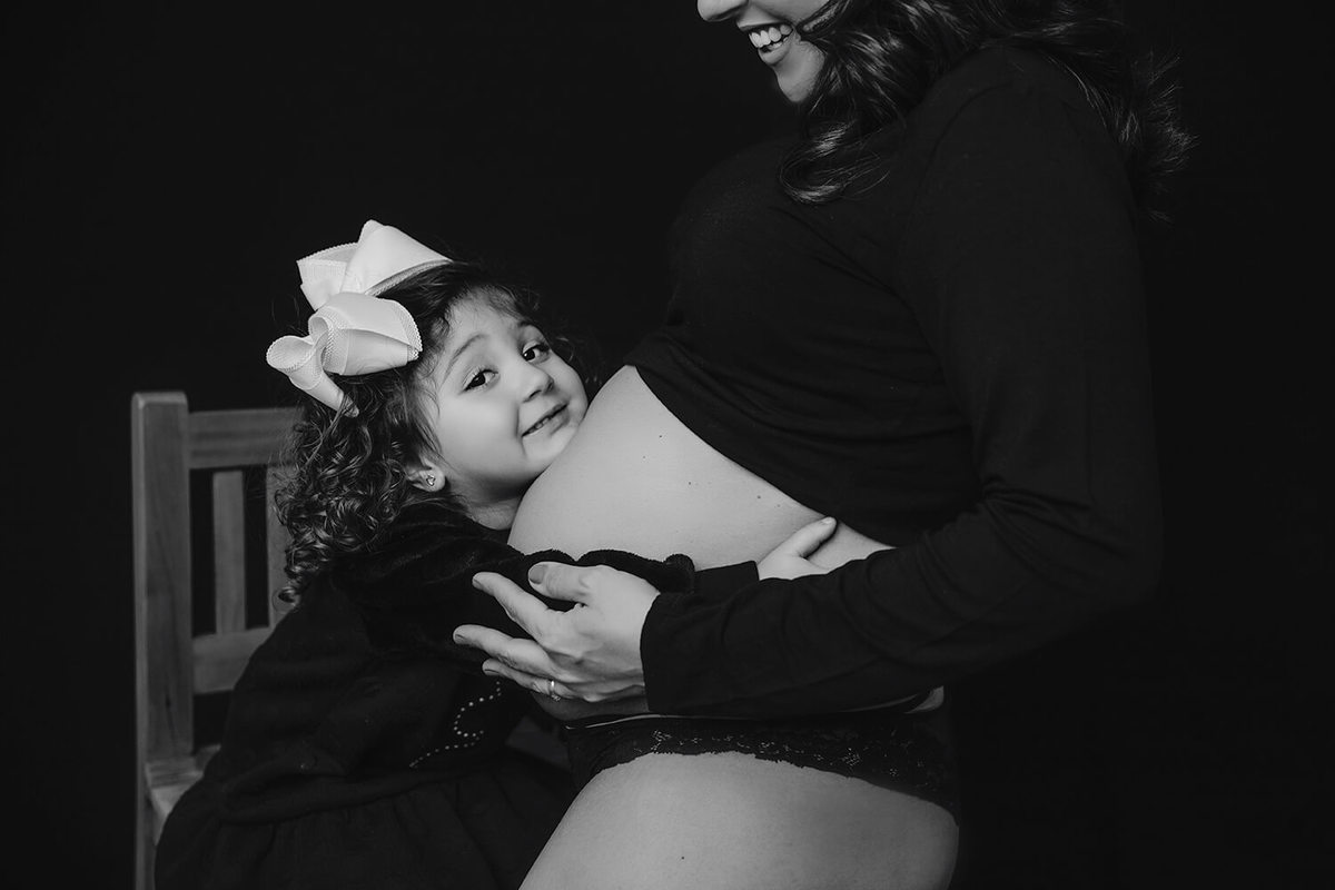 Gestante de lingerie preta com a mão na barriga e filhas abraçada no estúdio Natália Melo - Fotografia