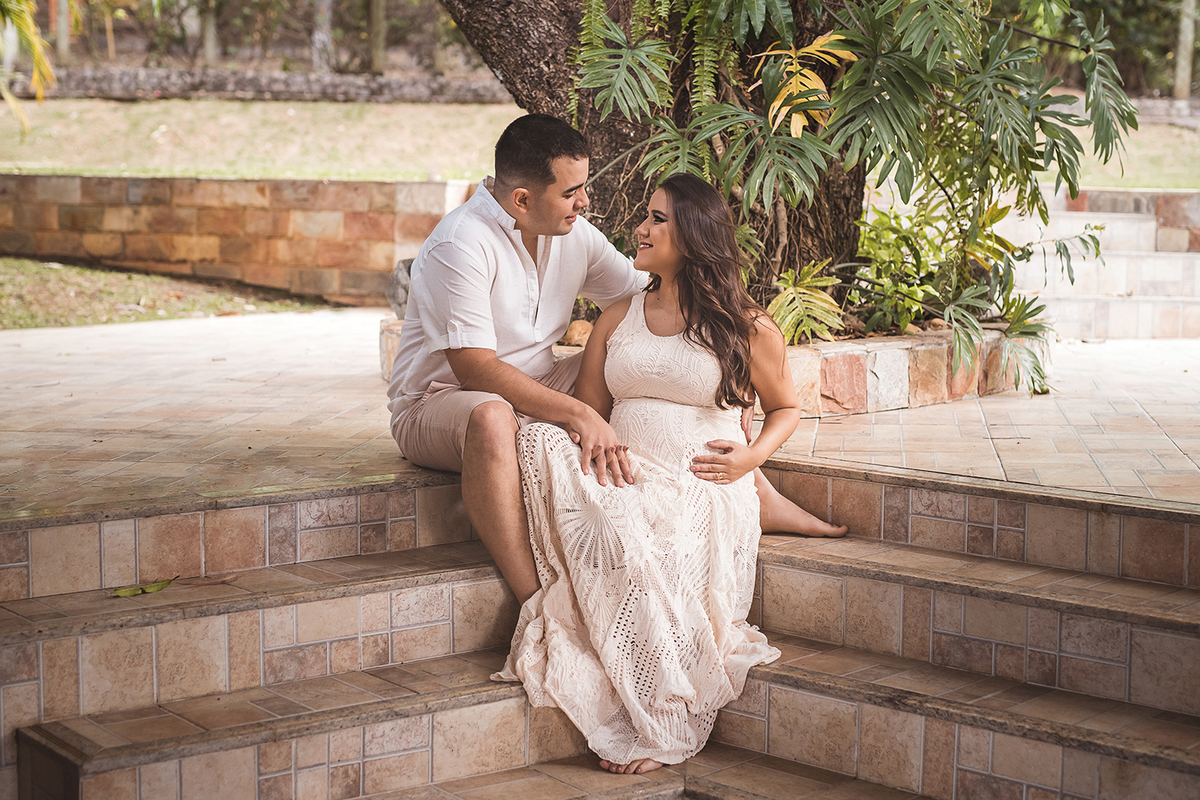 Gestante e esposo com roupa branca sentado numa escada olhando um para o outro em Belo Horizonte - MG