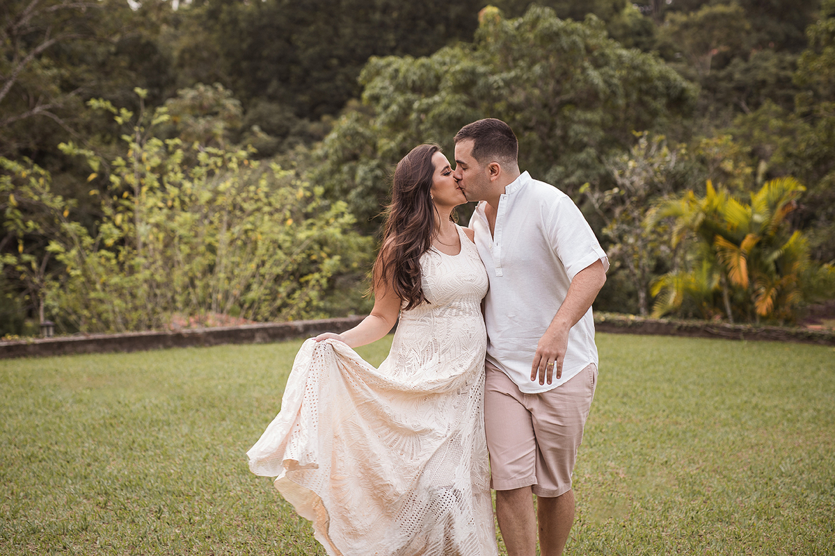 Gestante e esposo com roupa branca se beijando num campo gramado em Belo Horizonte - MG