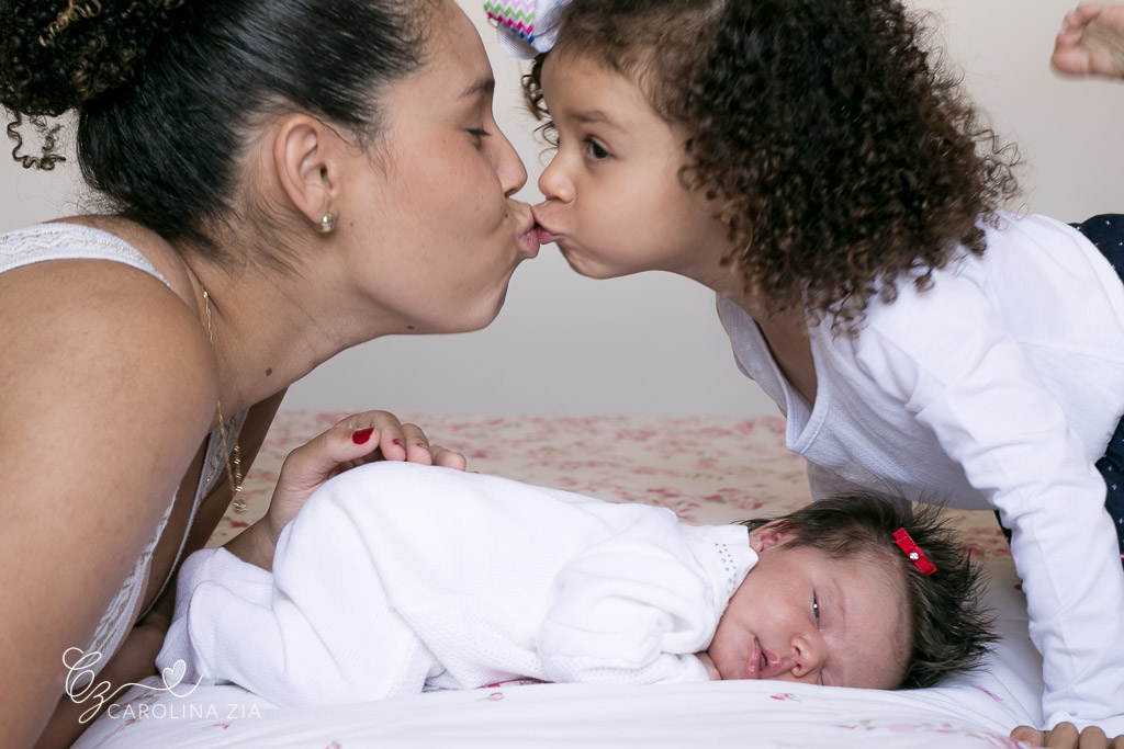 Mãe e filha se beijam enquanto bebê recém nascida dorme em sua sessão newborn