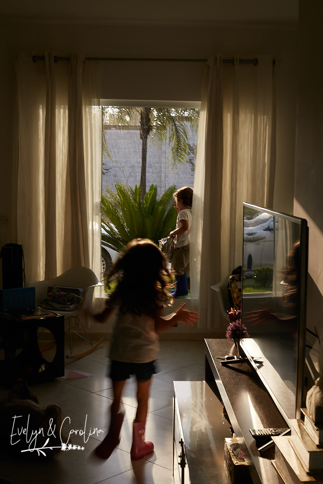 menina corre em direção ao irmãos que observa a chuva pela janela.