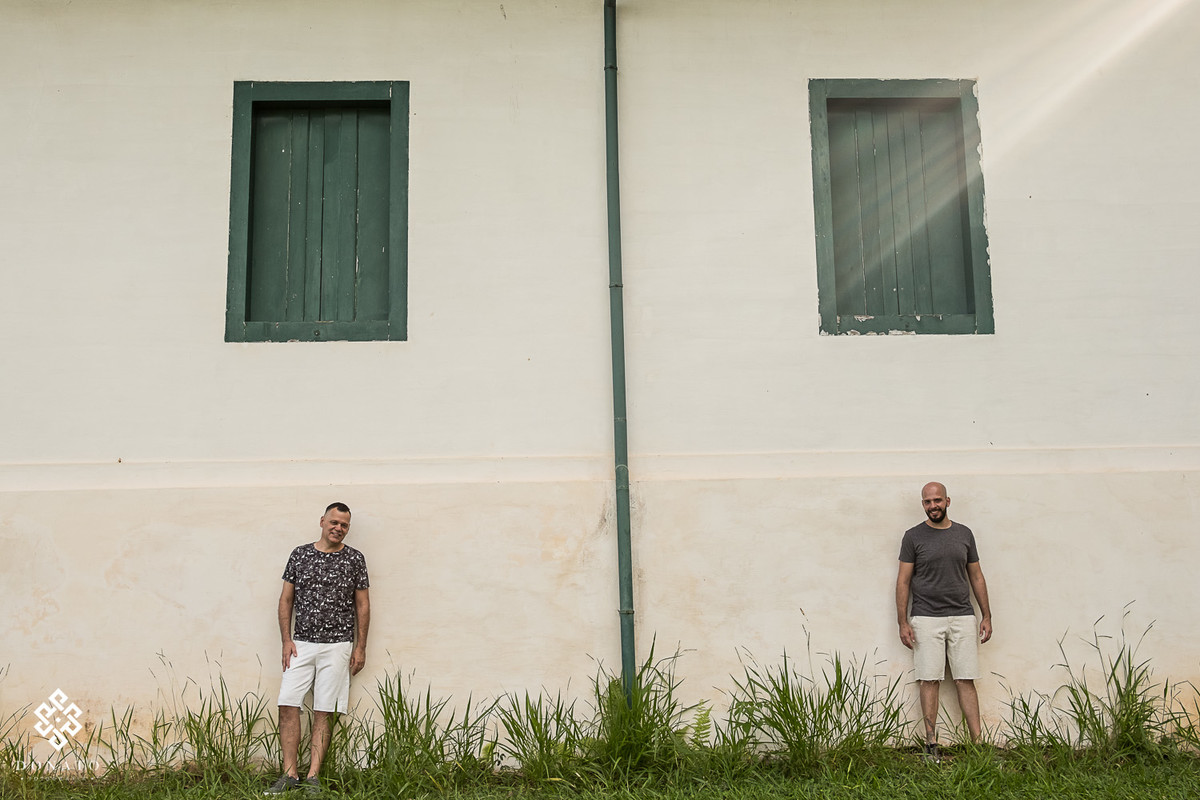 os garotos estam encostados em uma parede branca, acima tem duas janelas verde que faz uma composição muito interesante.