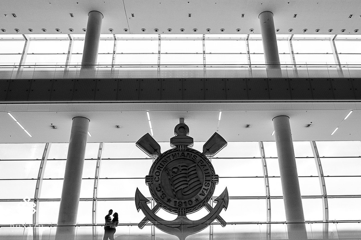Foto dos noivos dentro da arena Corinthians, com o símbolo atrás.