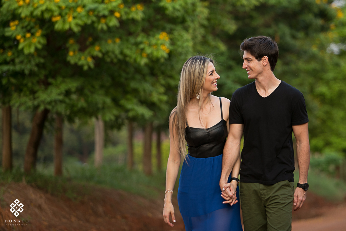 casal caminha em uma estrada de terra, o contraste do verde das arvores, com o marrom da terra, deixa esta imagem ainda mais bela.