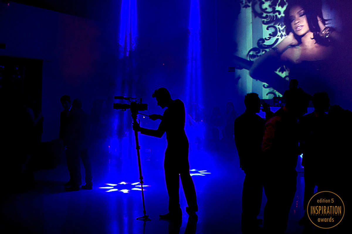 Foto do camera trabalhando com sua camera, a imagem é uma silhueta toda preta em meio a um fundo muito azul das luzes da balada, a foto teve o premio no inspirations em 2014.