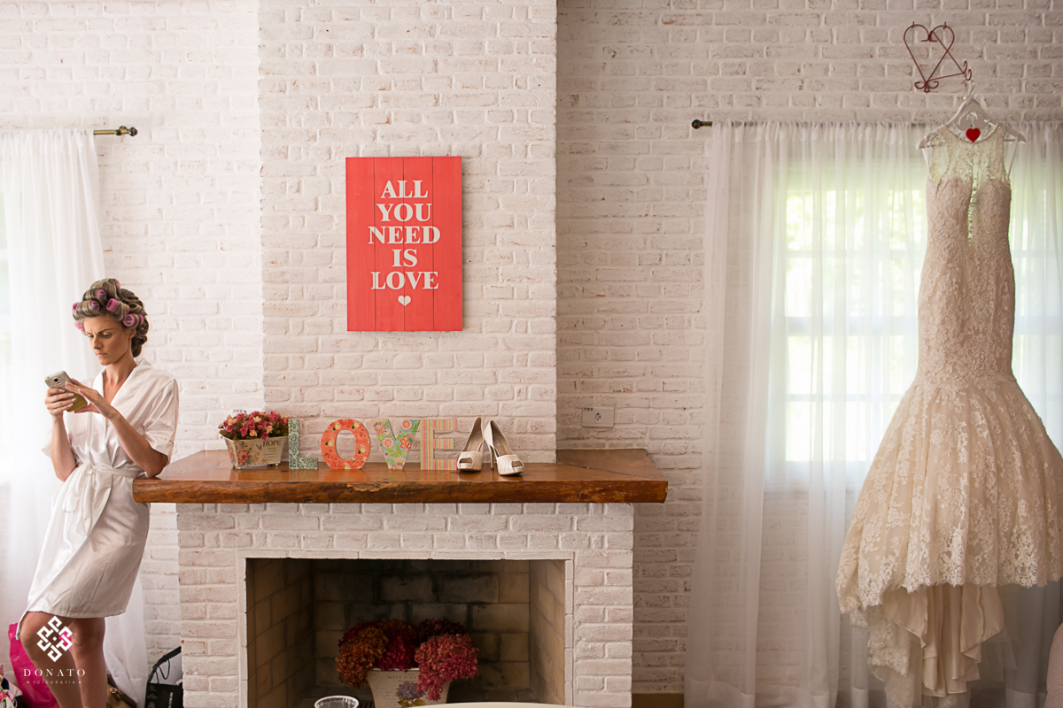detalhe da sala do making da noiva, onde tem o detalhe do vestido ao lado direito e a noiva ao lado esquerdo com o celular na mão, ao meio tem uma placa dizendo All You Need is Love.